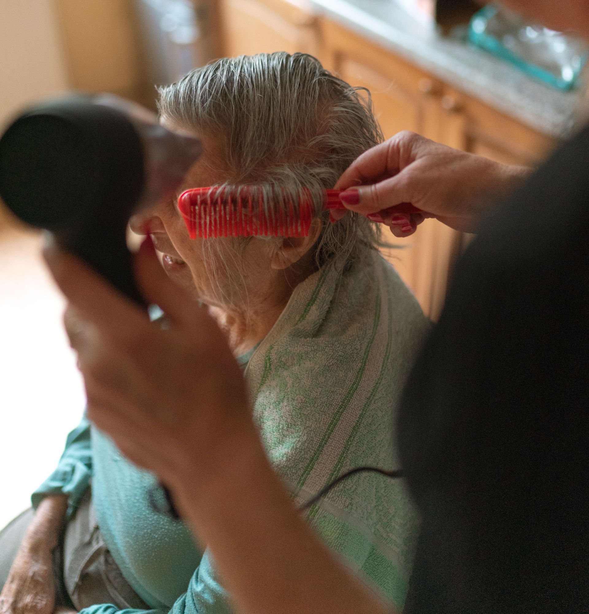 A caregiver assists a senior adult with hair drying and combing at home.